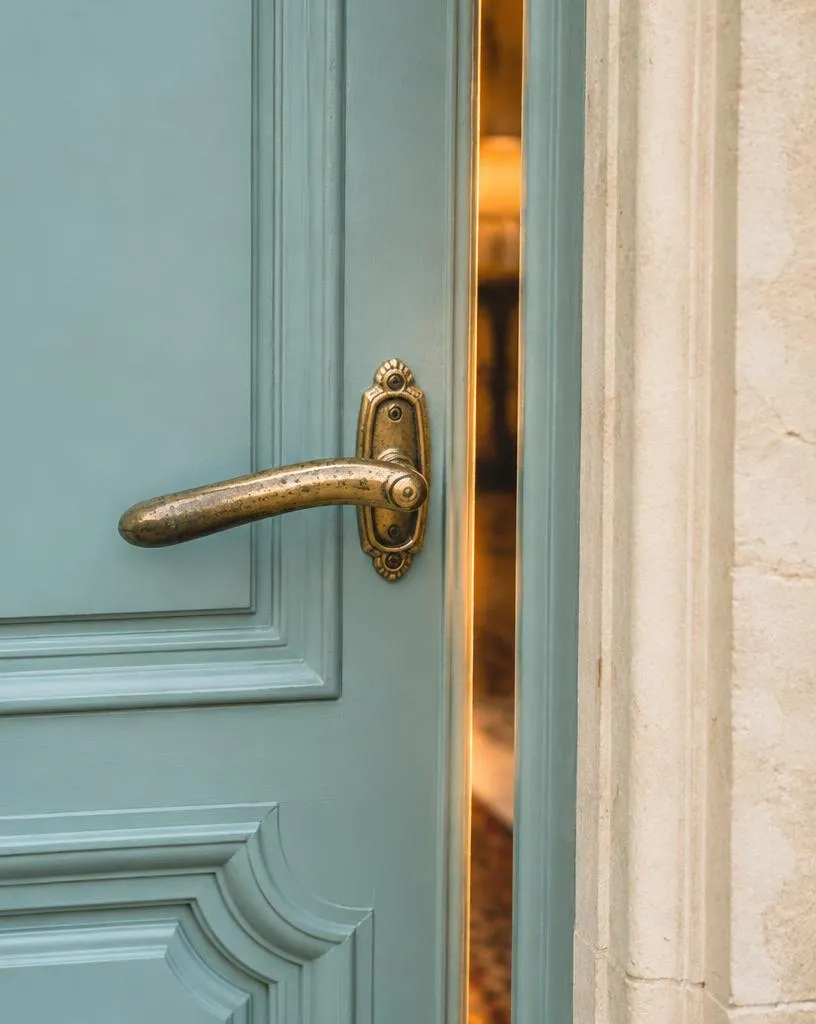 Aged brass handle on a partially open robin's-egg-blue Parisian door with warm light in the gap